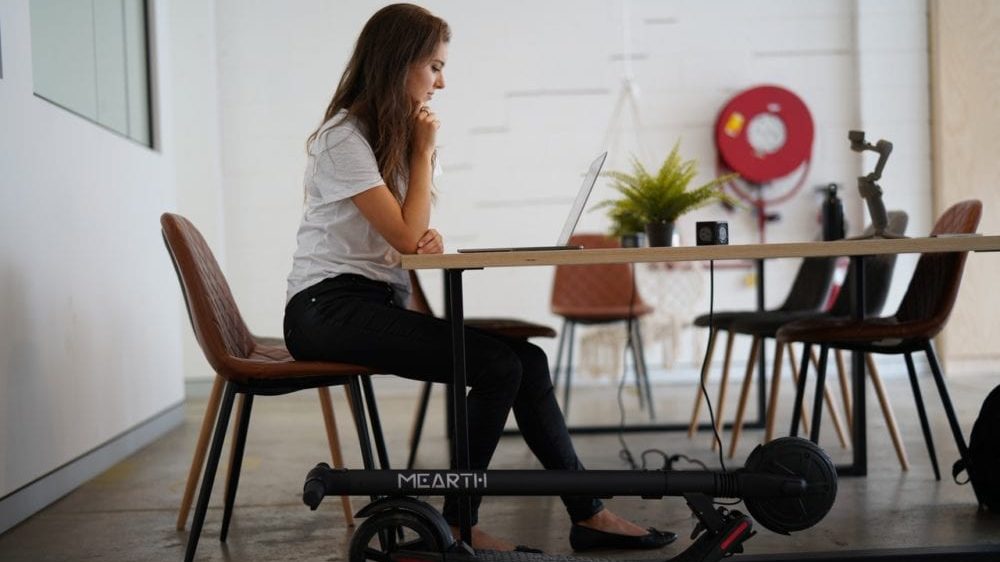 Zijaanzicht van een vrouw aan tafel, met opgevouwen e-scooter naast haar.