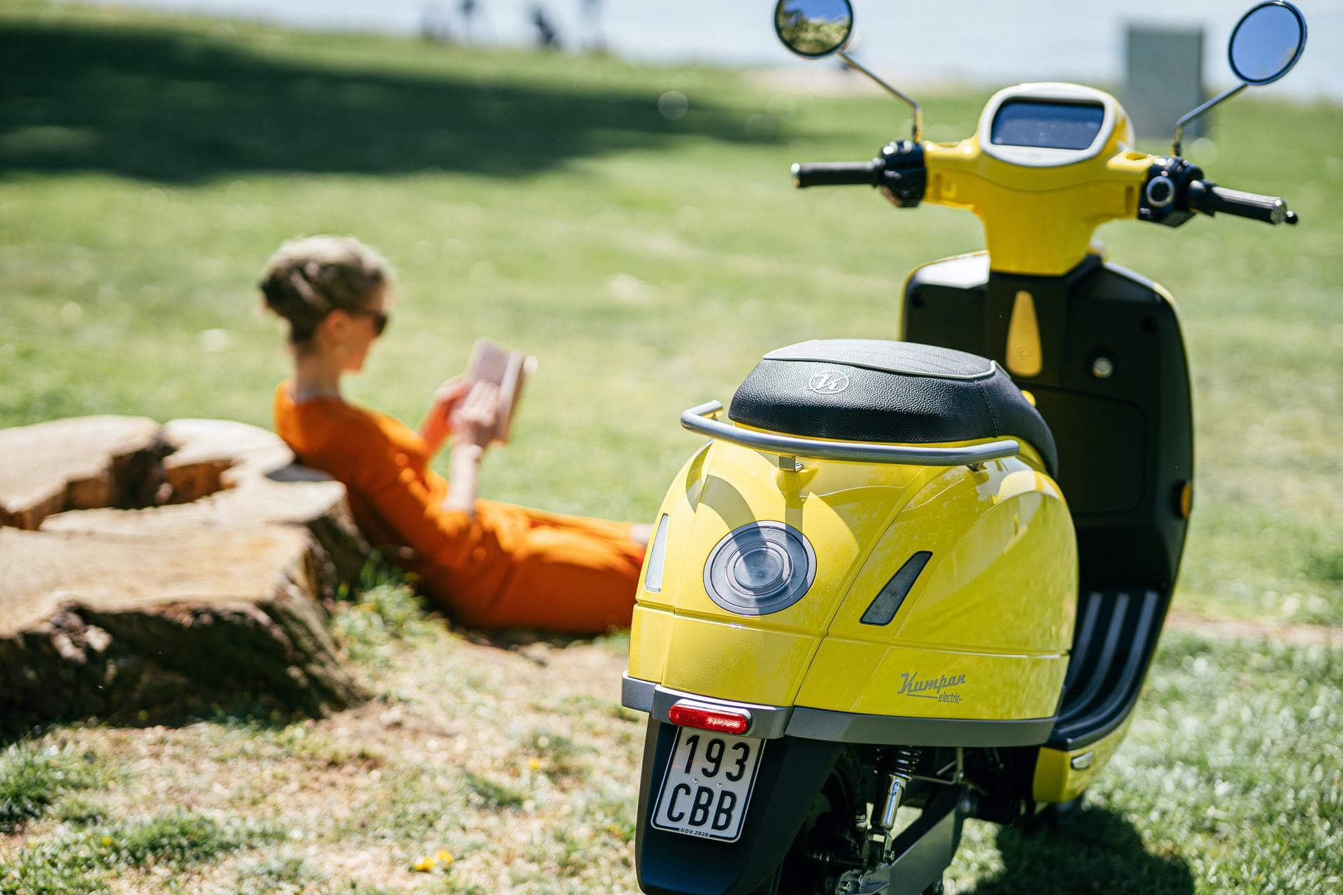 Gele scooter van achteren op een veld bij iemand die tegen een steen aan zit